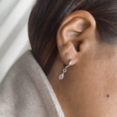 עגיל חישוק כסף אמיתי 925 עם תליון בצורת טיפה משובץ זרקון, על אוזן אישה עם שיער חום ואאוטפיט בגוון בז