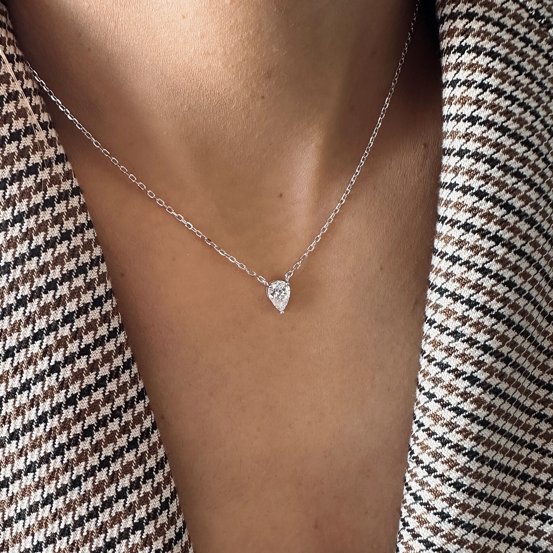 שרשרת כסף דקה עם תליון בצורת טיפה משובץ אבן נוצצת, מוצגת על צוואר אישה הלובשת ז’קט טוויד בגווני חום ולבן.