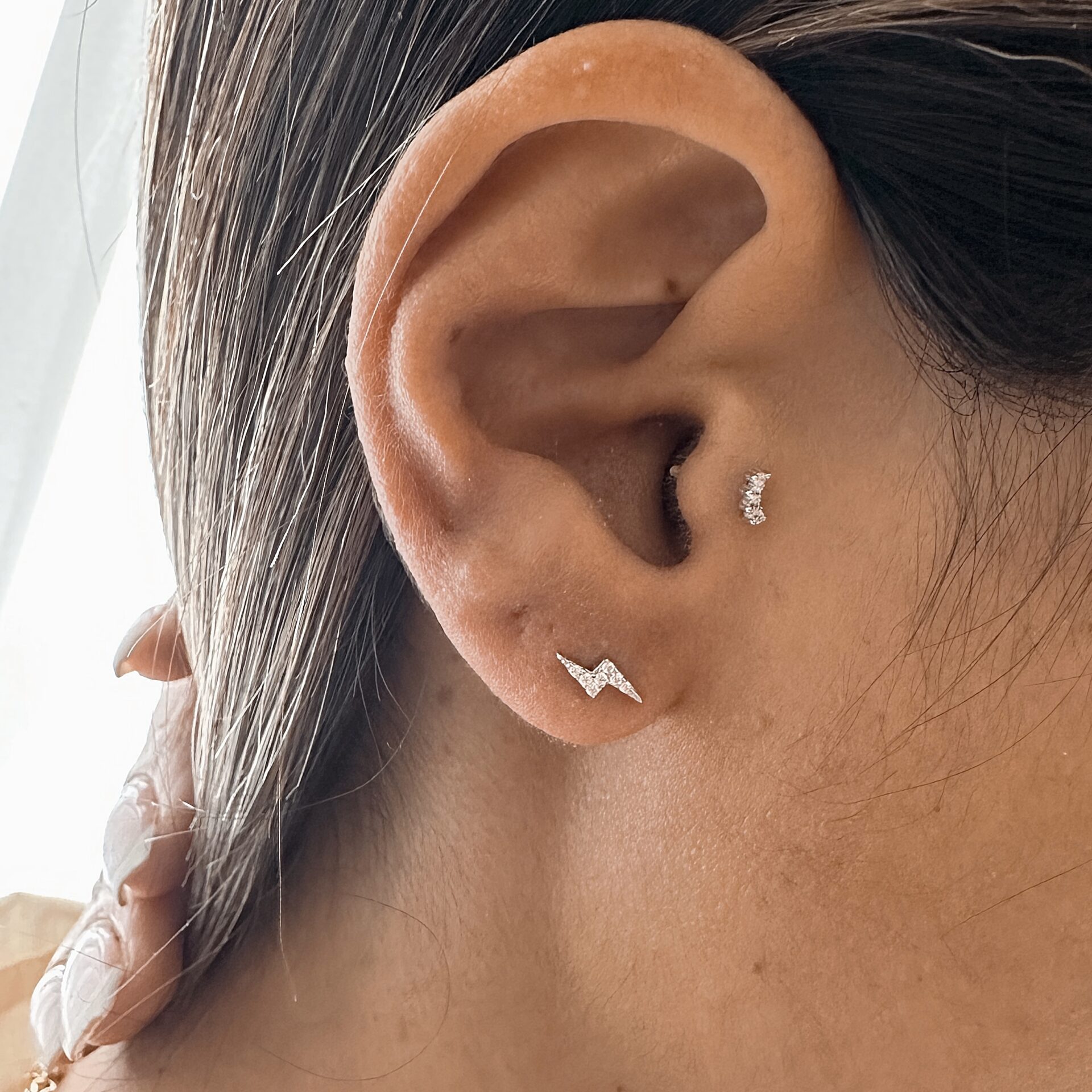 אוזן של אישה עם שני עגילי כסף 925 משובצים בזירקונים – עגיל קטן בצורת ברק בתנוך האוזן ועגיל קטן עם שלושה זירקונים בחלק העליון של האוזן.כסף אמיתי 925