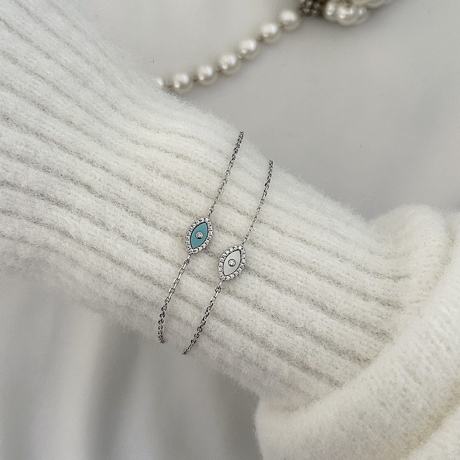 צמידי עין צדף כסף 925 משובצים זרקונים, אחד עם אבן כחולה ואחד עם אבן לבנה, מונחים על סוודר לבן סמוך לשרשרת פנינים