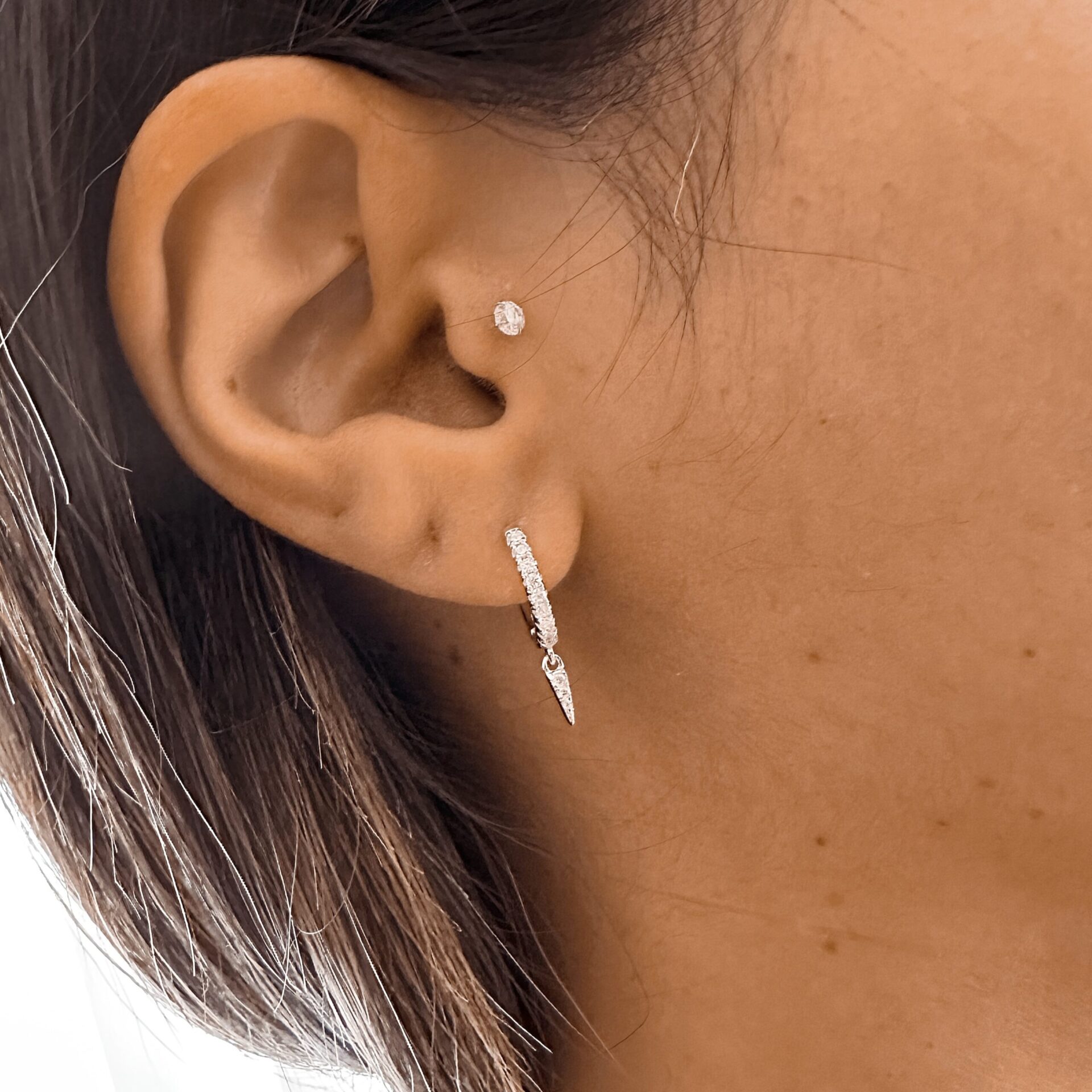 עגיל חישוק כסף 925 משובץ זרקונים עם תליון קטן בצורת שן, מצולם על אוזן של אישה עם שיער חום כהה ופירסינג נוסף עם אבן קטנה