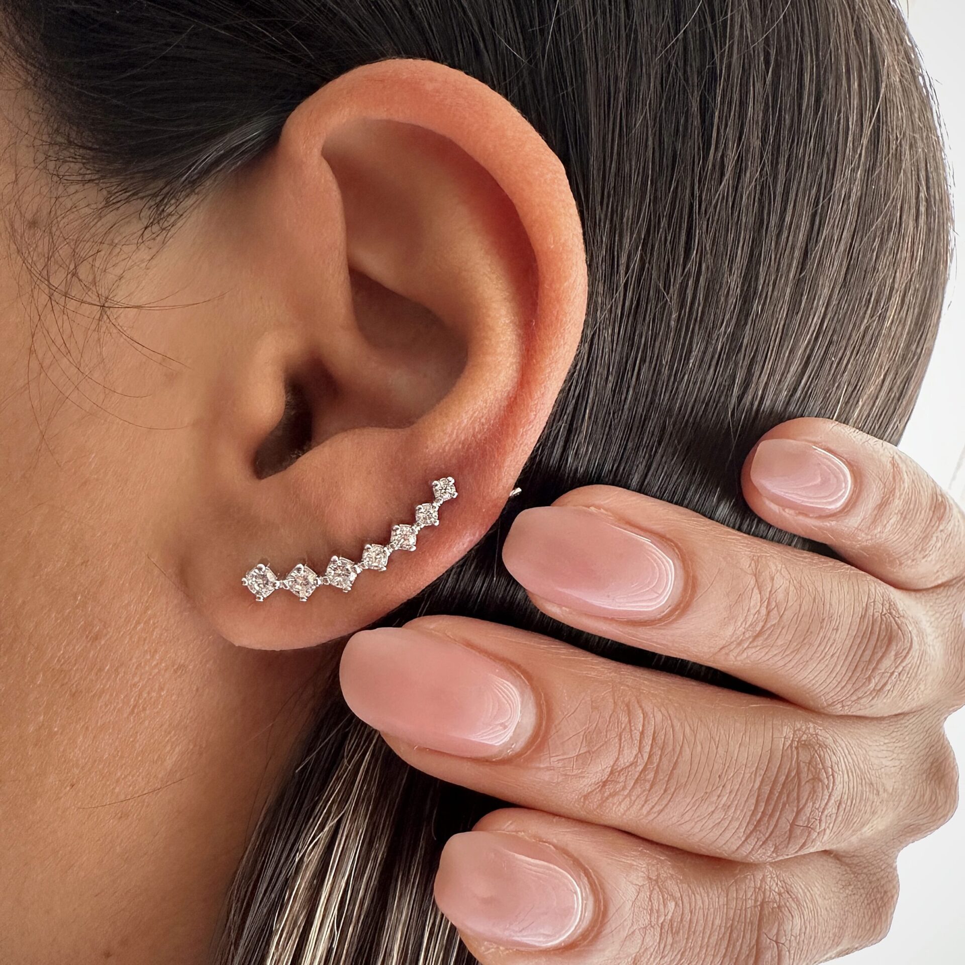 עגיל מטפס מעוצב בצורת שורת יהלומים קטנים, משובץ זרקונים, מוצמד לאורך תנוך האוזן של אישה. ידה עם לק ורוד טבעי מונחת על שיערה החלק.