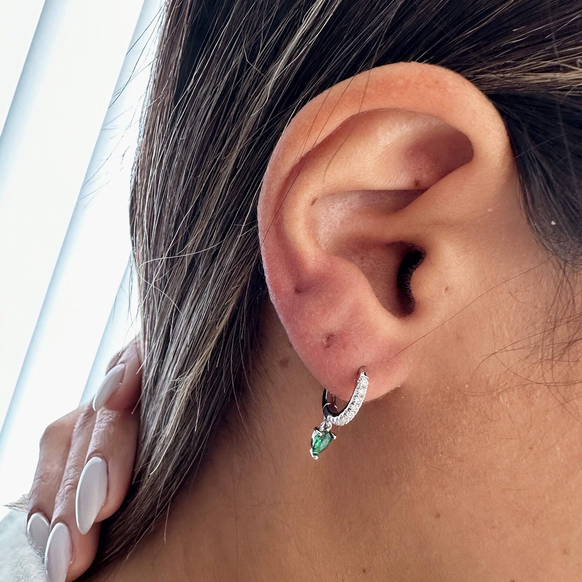 עגיל כסף קטן משובץ אבנים לבנות עם תליון טיפה ירוק, תלוי על תנוך האוזן של אישה עם שיער חום בהיר. היד עם ציפורניים מטופחות נוגעת בעורף. כסף אמיתי 925